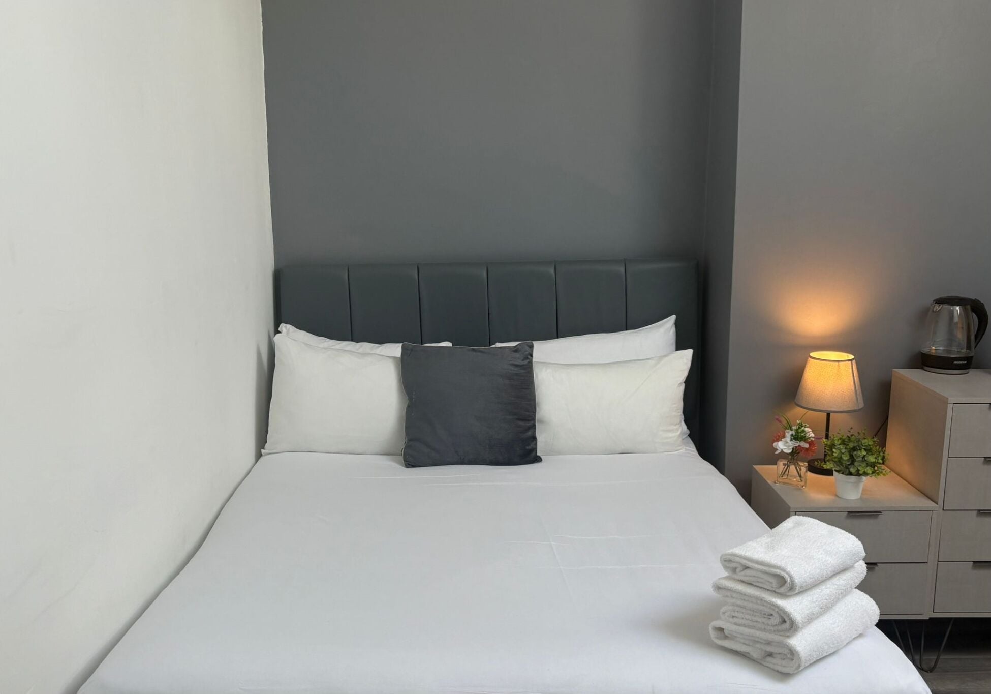A neatly made bed with white linens and a dark headboard, a bedside table with a lamp and a small plant, and a stack of folded towels.