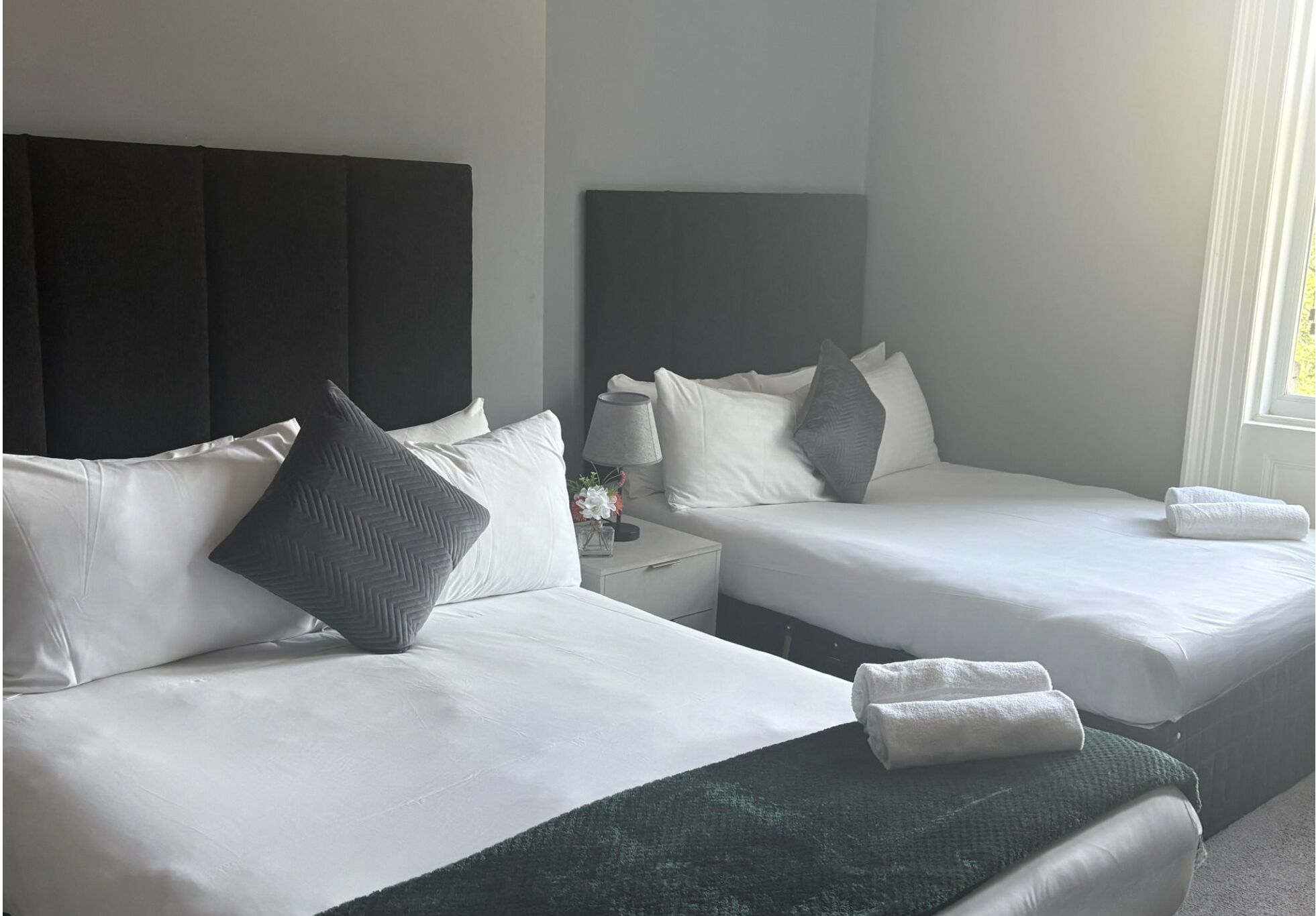 Una habitación de hotel con dos camas, cada una con ropa de cama y almohadas blancas, una mesita de noche con una lámpara y flores, y una ventana con luz natural.