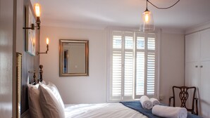 A bedroom with a bed, a chair, a window with shutters, and wall-mounted lights. - The Blacksmiths Arms (Lewes)