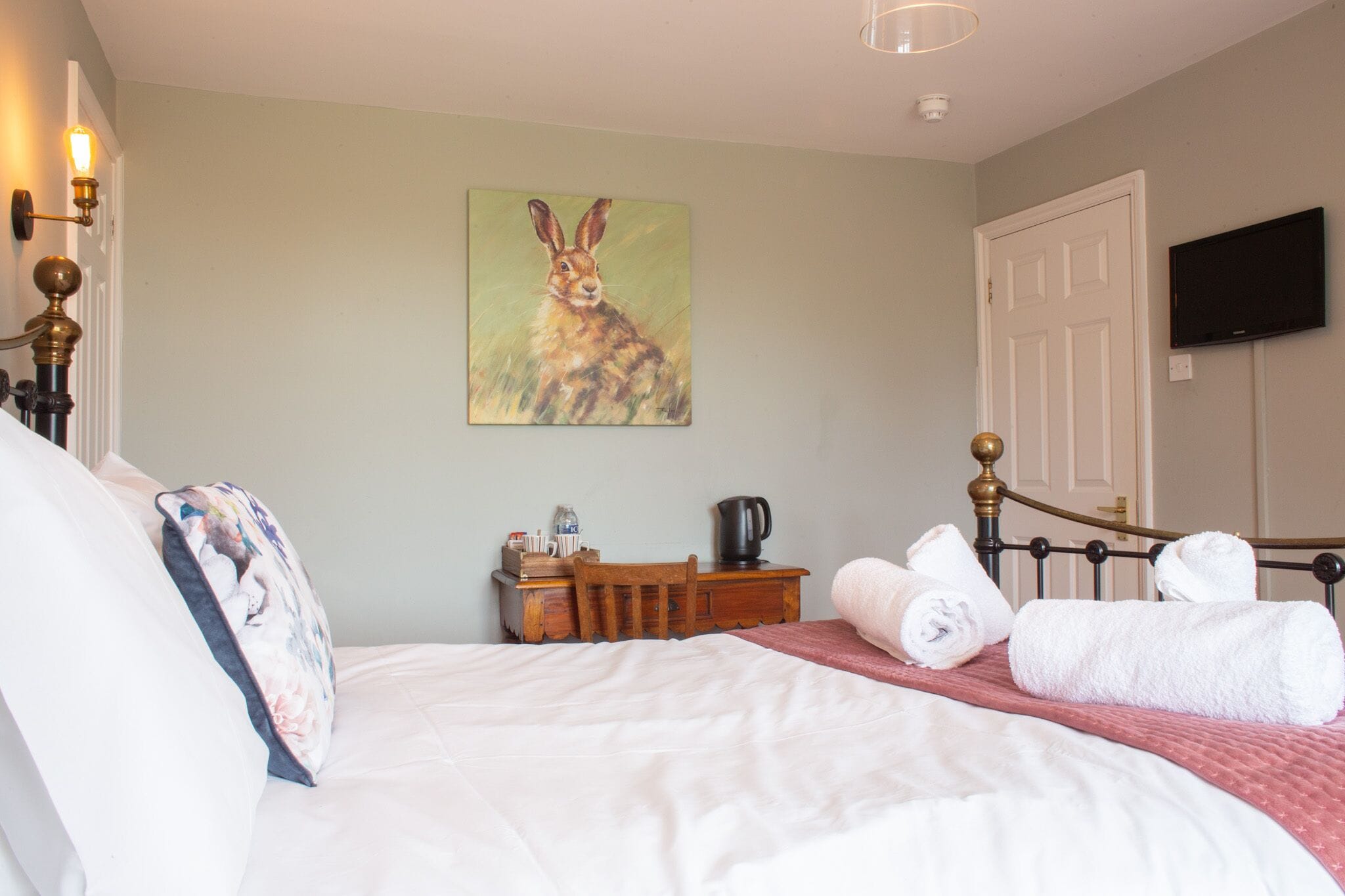 A neatly made bed with white linens, a wooden nightstand with a kettle, a painting of a rabbit on the wall, and a television mounted on the wall.