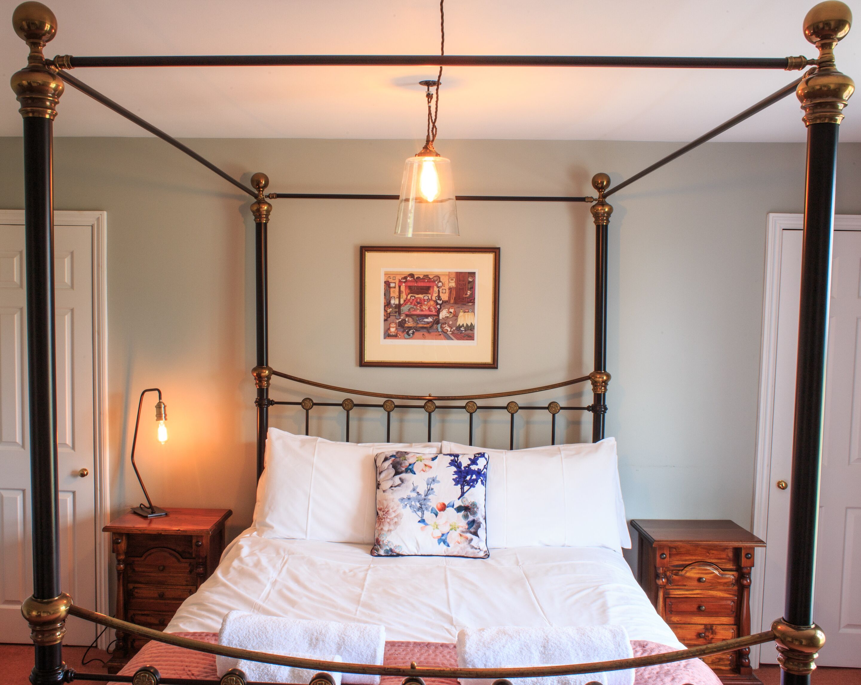 A four-poster bed with a white bedspread and a floral pillow, a bedside table, a nightstand, and a framed picture on the wall.