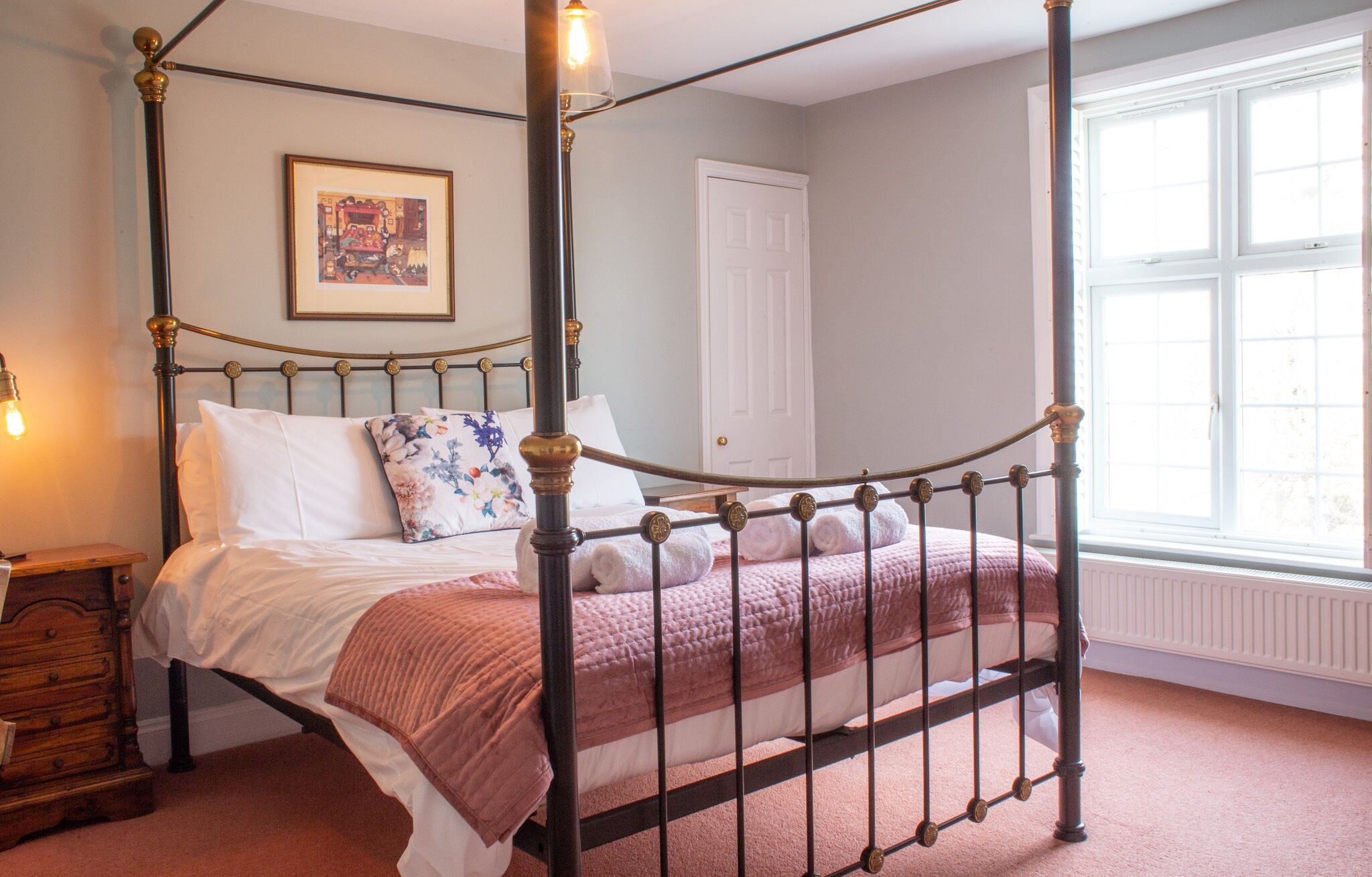 A four-poster bed with rolled towels, a bedside table with a lamp, a framed picture on the wall, and a window with blinds.