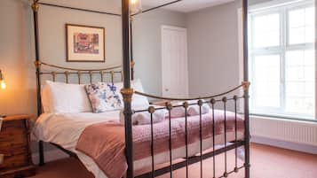 A four-poster bed with rolled towels, a bedside table with a lamp, a framed picture on the wall, and a window with blinds.