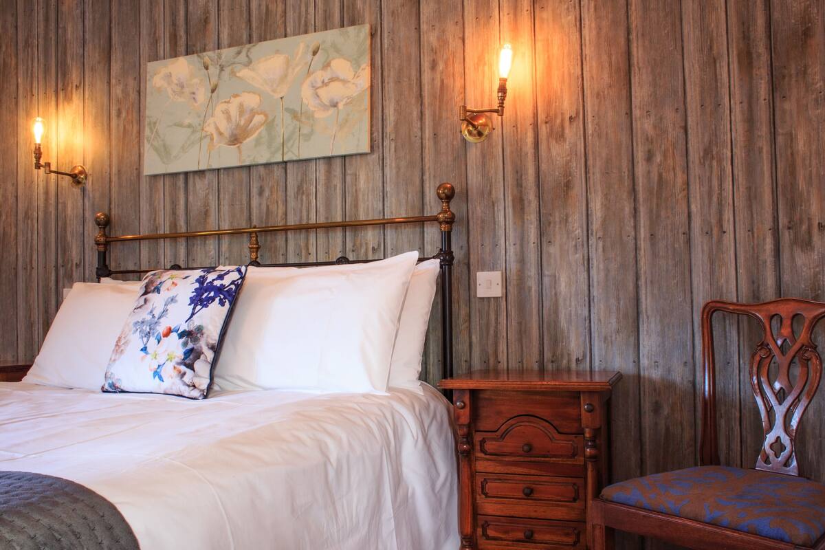 Une pièce aux murs lambrés, comprenant un lit, une table de chevet en bois, une chaise et des lampes fixées au mur.