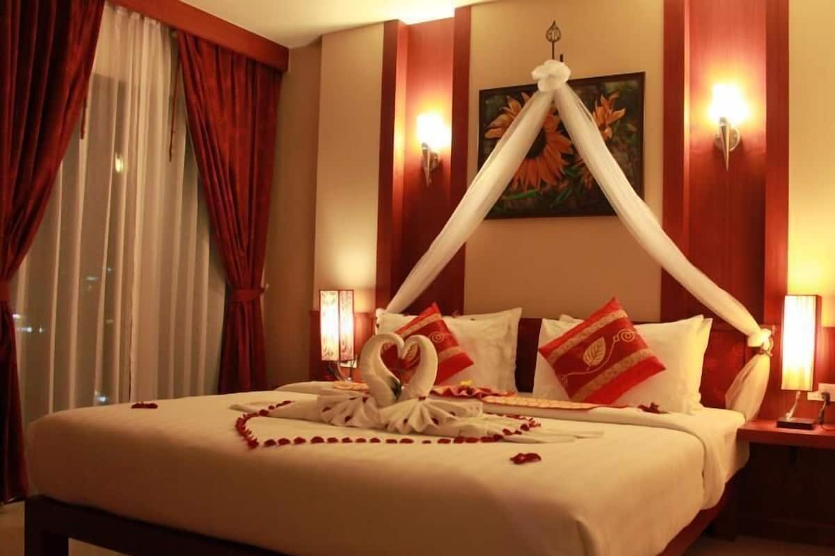 A hotel room with a bed, red curtains, a painting, and wall sconces.