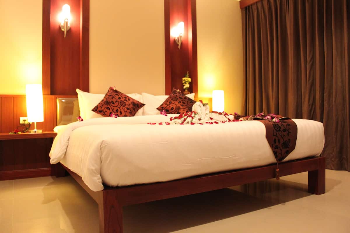 A neatly made bed with white linens and decorative pillows, flanked by bedside tables with lamps, in a room with wooden paneling and a large window with curtains.