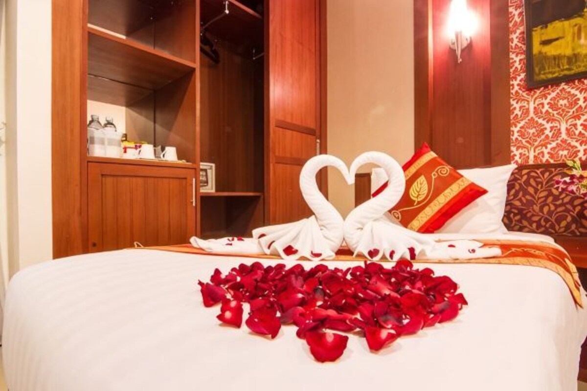 Ein Hotelzimmer mit einem ordentlich bezogenen Bett, einer herzförmigen Handtuchgestaltung und einer mit Rosenblättern bedeckten Bettdecke.