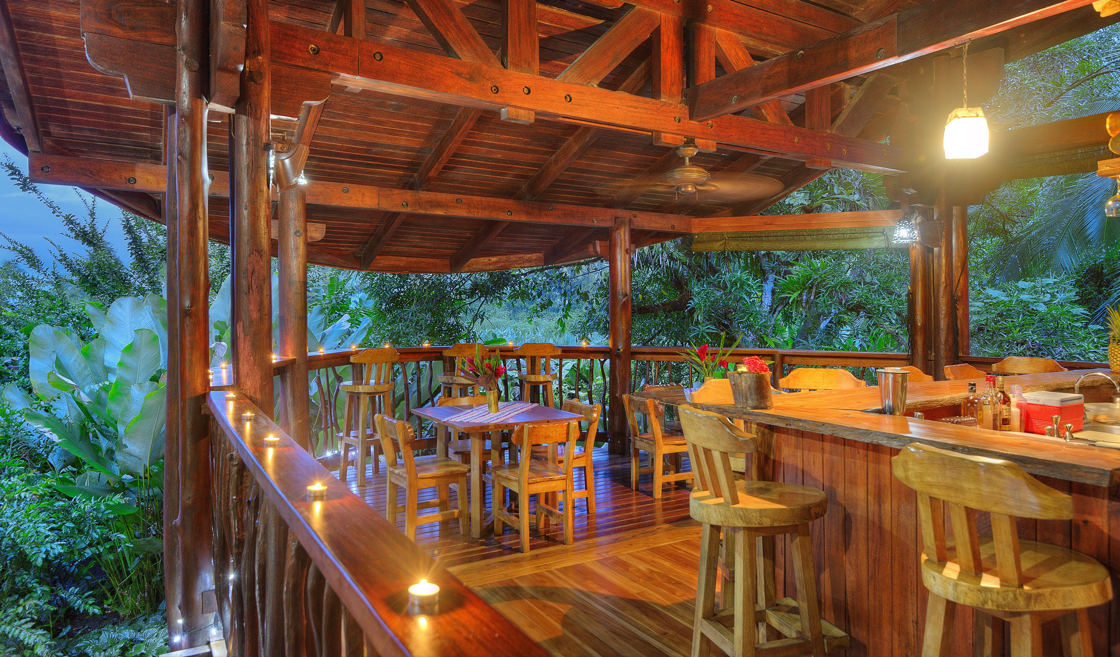 Une terrasse extérieure en bois, agrémentée de tables et de chaises, et entourée d’une végétation luxuriante.