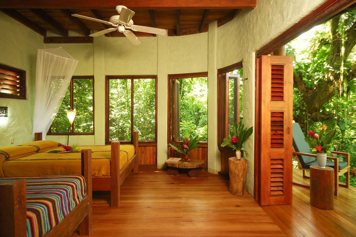 A room with wooden flooring, a bed with a colorful striped bedspread, a ceiling fan, and large windows offering a view of greenery outside.