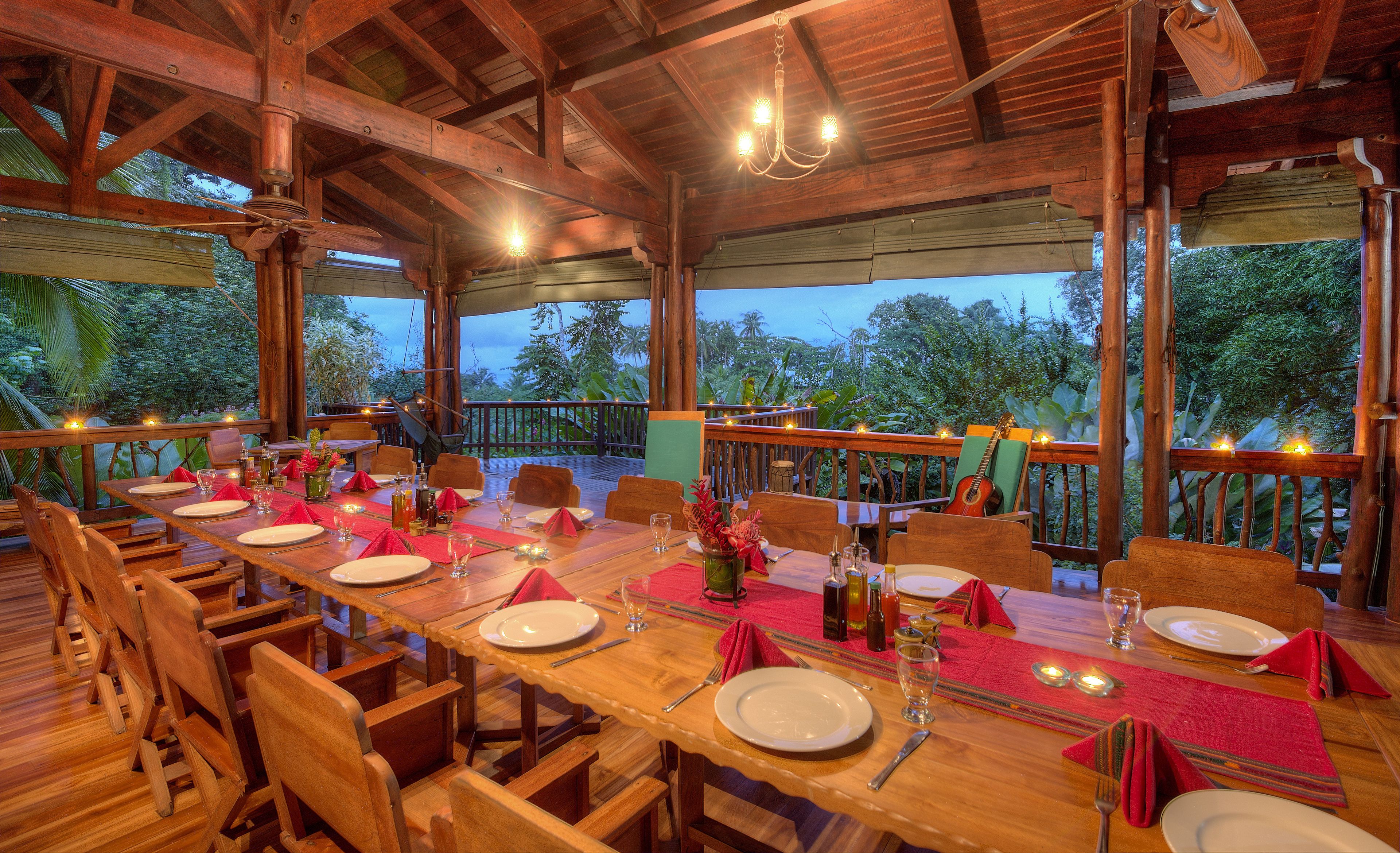A wooden dining area with tables set for a meal, surrounded by wooden railings and overlooking a lush garden.