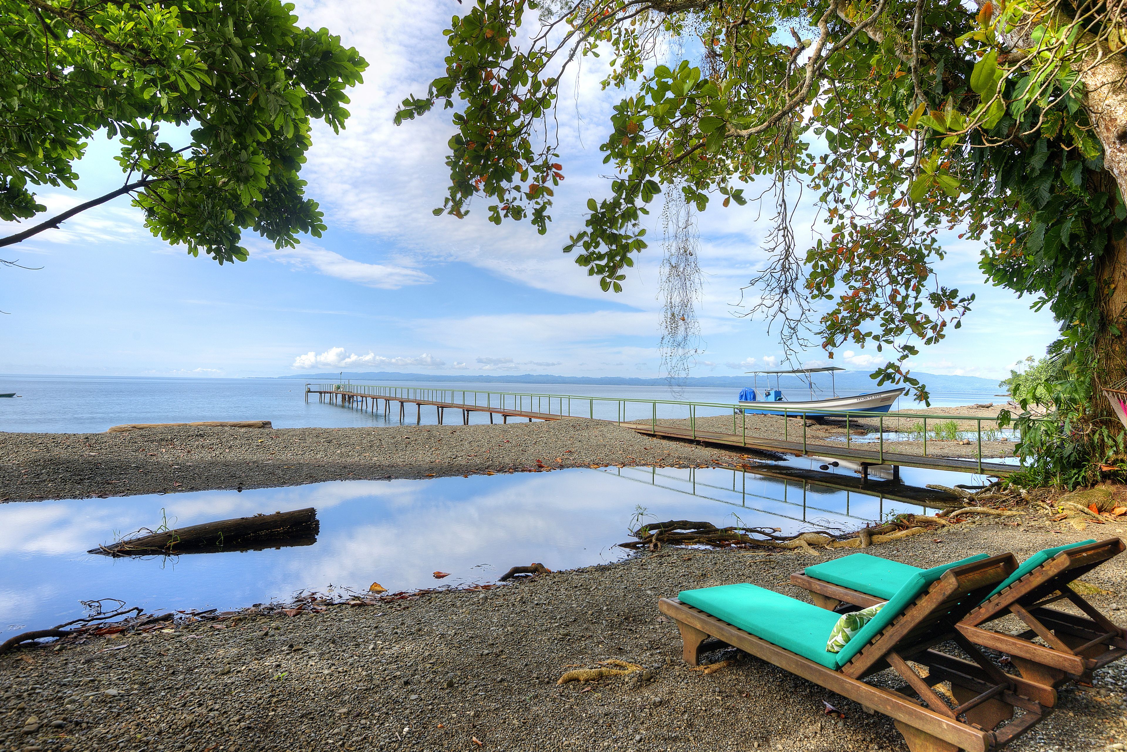 A beach with wooden lounge chairs, a wooden pier extending into calm waters, and a boat docked at the end.