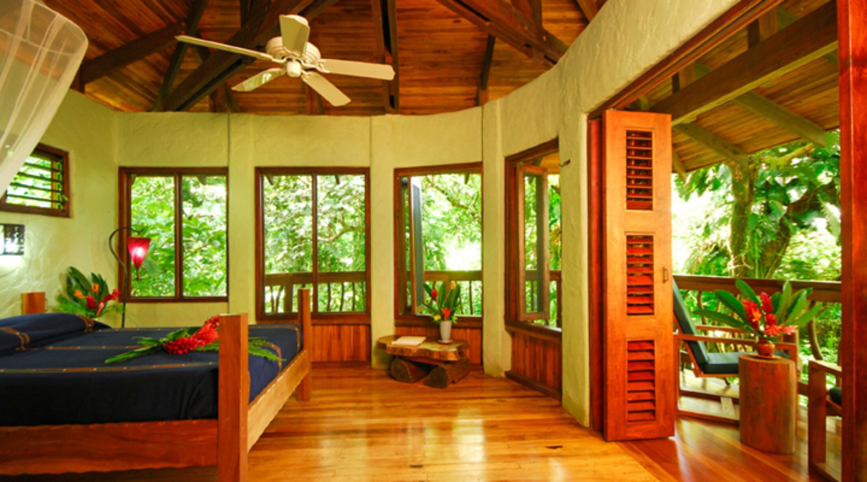 A room with a bed, a ceiling fan, large windows, and a view of the outdoors.