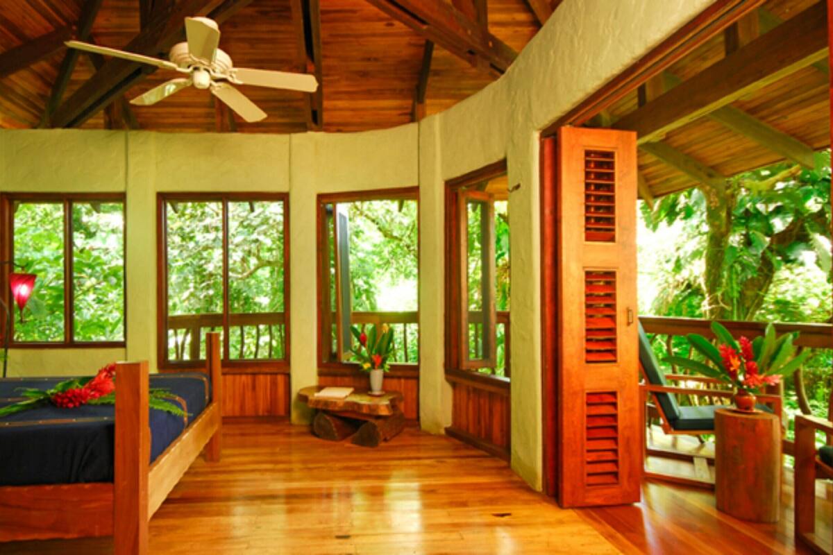 A room with a bed, a ceiling fan, large windows, and a view of the outdoors.