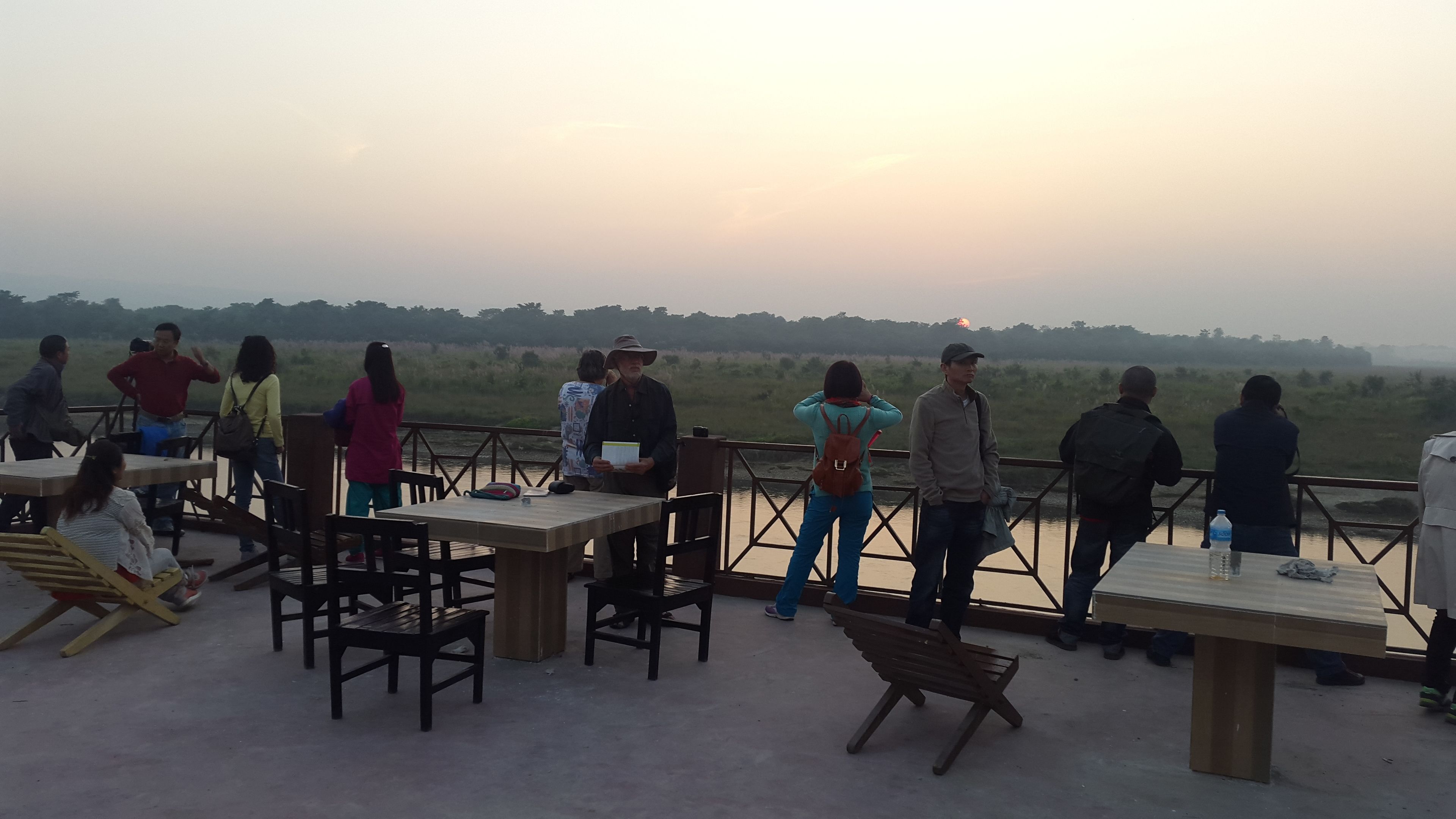 A group of people sitting at tables on an outdoor terrace with a view of a river and distant hills.