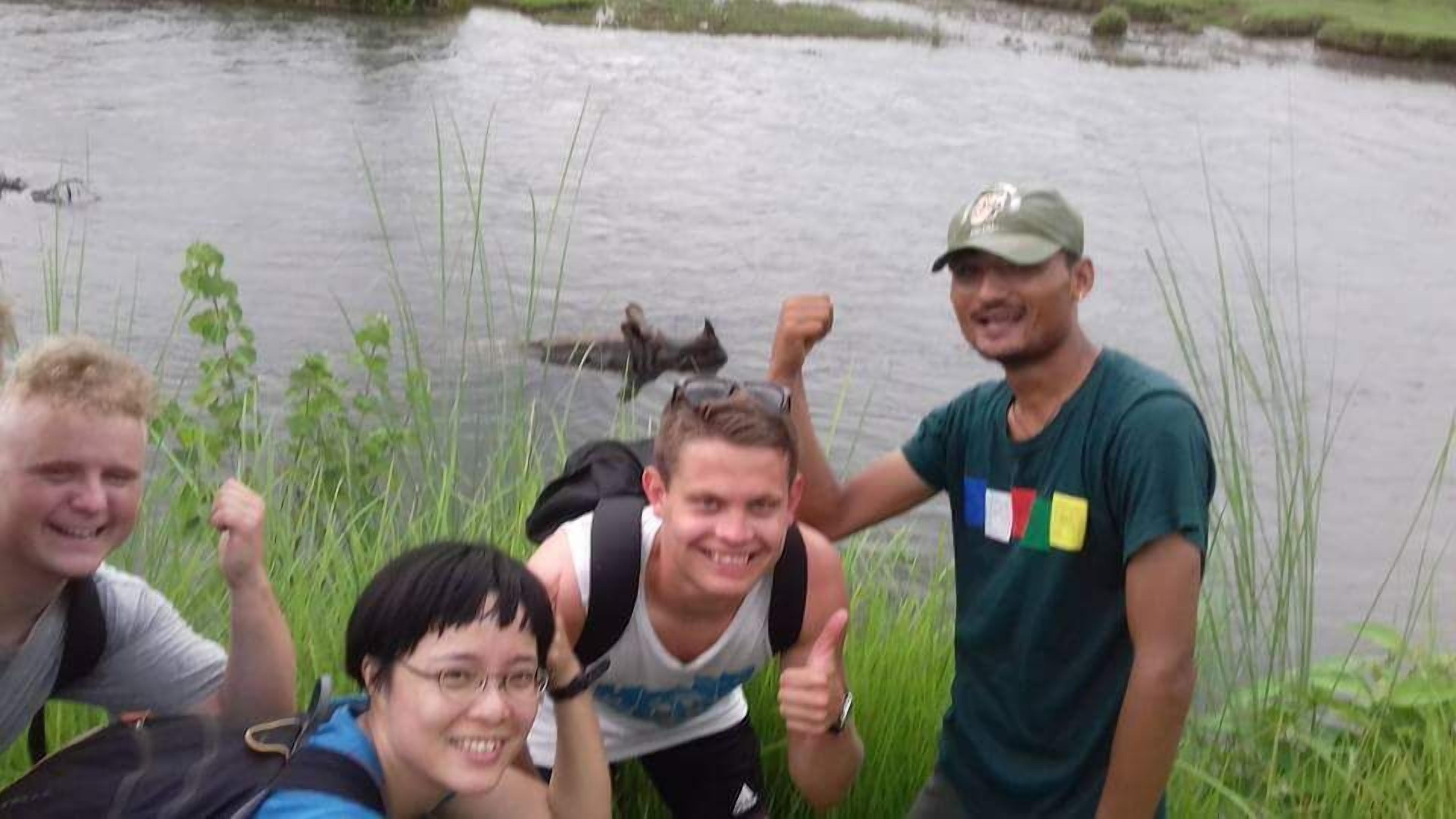 Un groupe de personnes se trouve près d’une étendue d’eau calme, peut-être un lac ou une rivière, avec de hautes herbes et des roseaux au premier plan.
