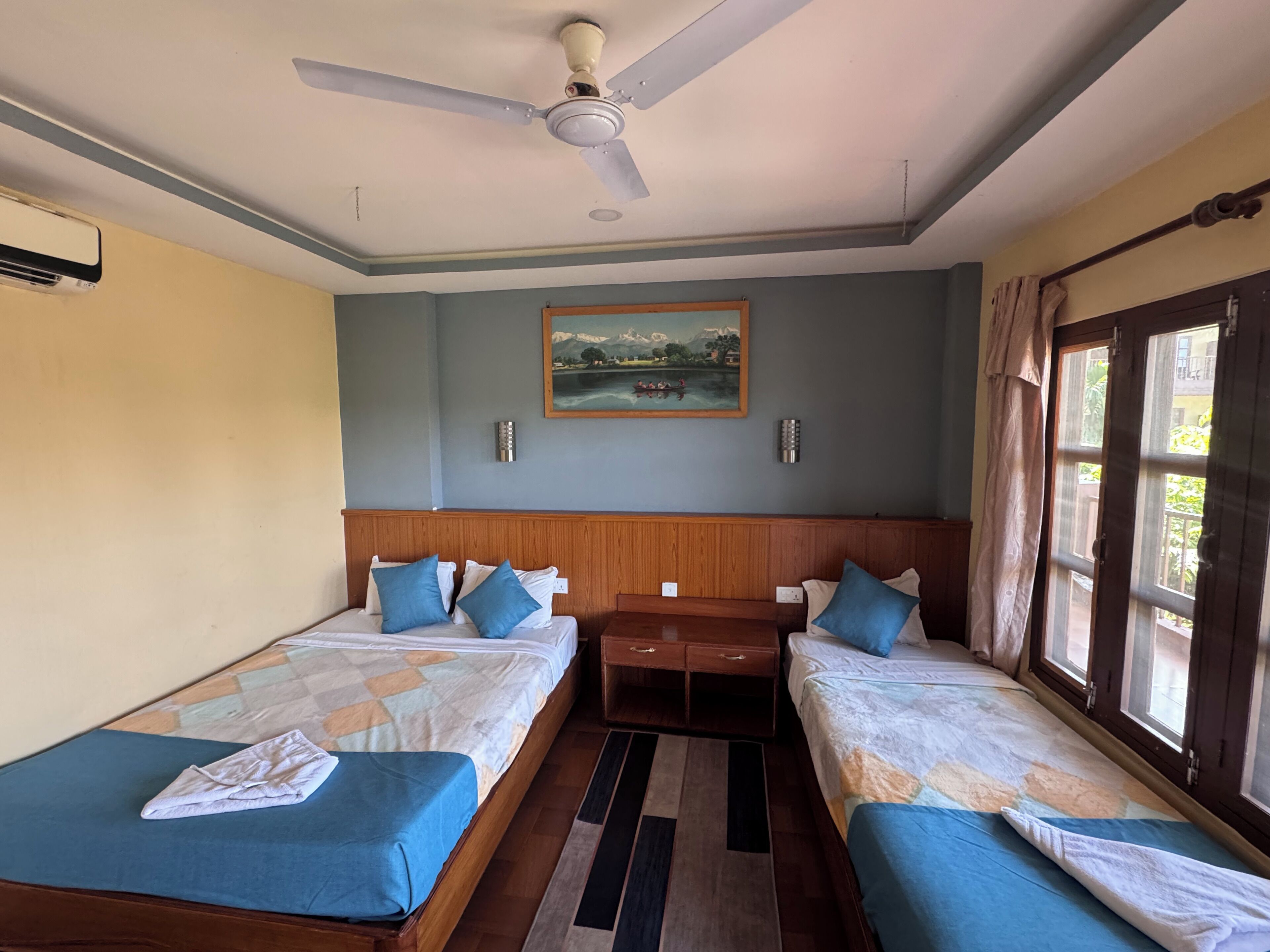 Una camera d'albergo con due letti, una ventola a soffitto, un quadro appeso e una finestra con tende.