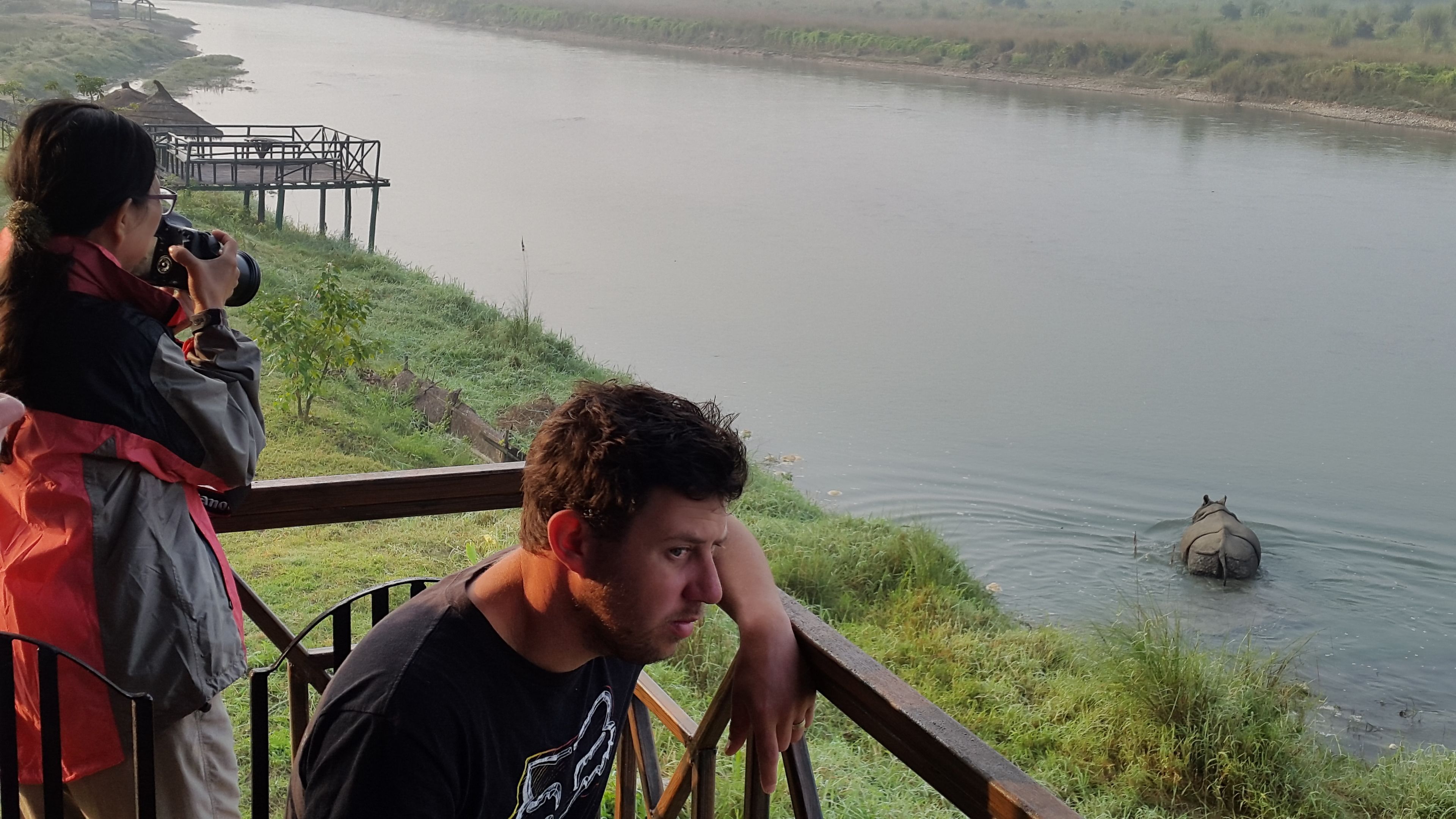 A person sitting on a wooden deck overlooking a river, with another person taking a photo.