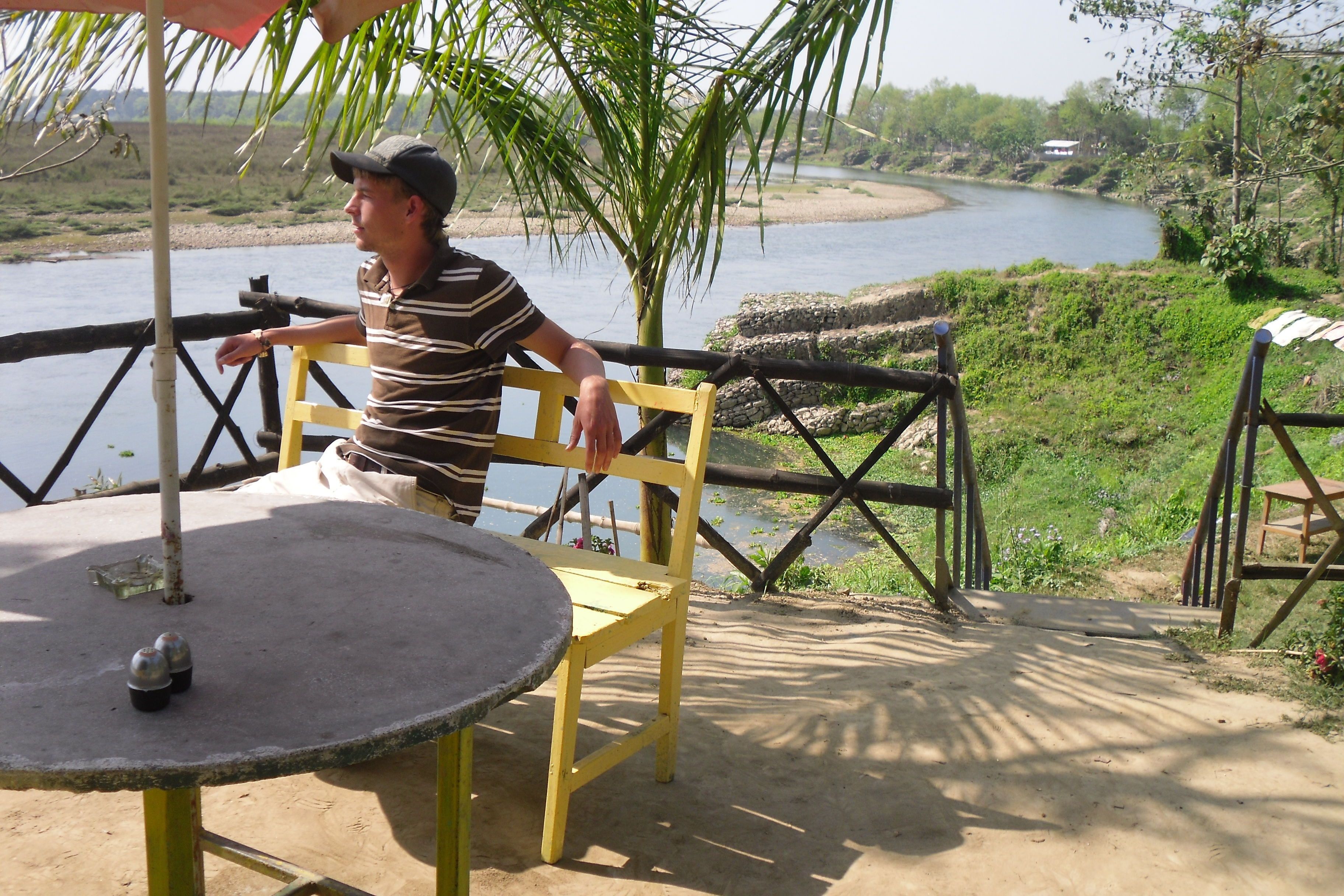 A man sitting on a yellow chair at a table with a metal top, overlooking a river.