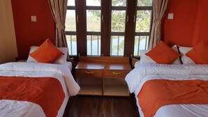 A hotel room with two beds, each with white bedding and orange accents. There is a wooden nightstand between the beds, and the room has large windows with curtains. - Jungle Wildlife Camp (Sauraha)