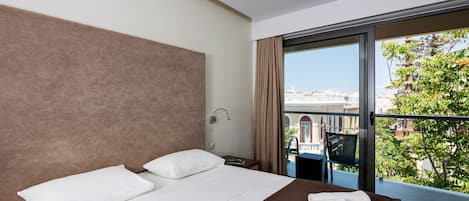 Habitación de hotel con una cama grande, una mesita de noche, una lámpara y vistas a la ciudad a través de un ventanal amplio.