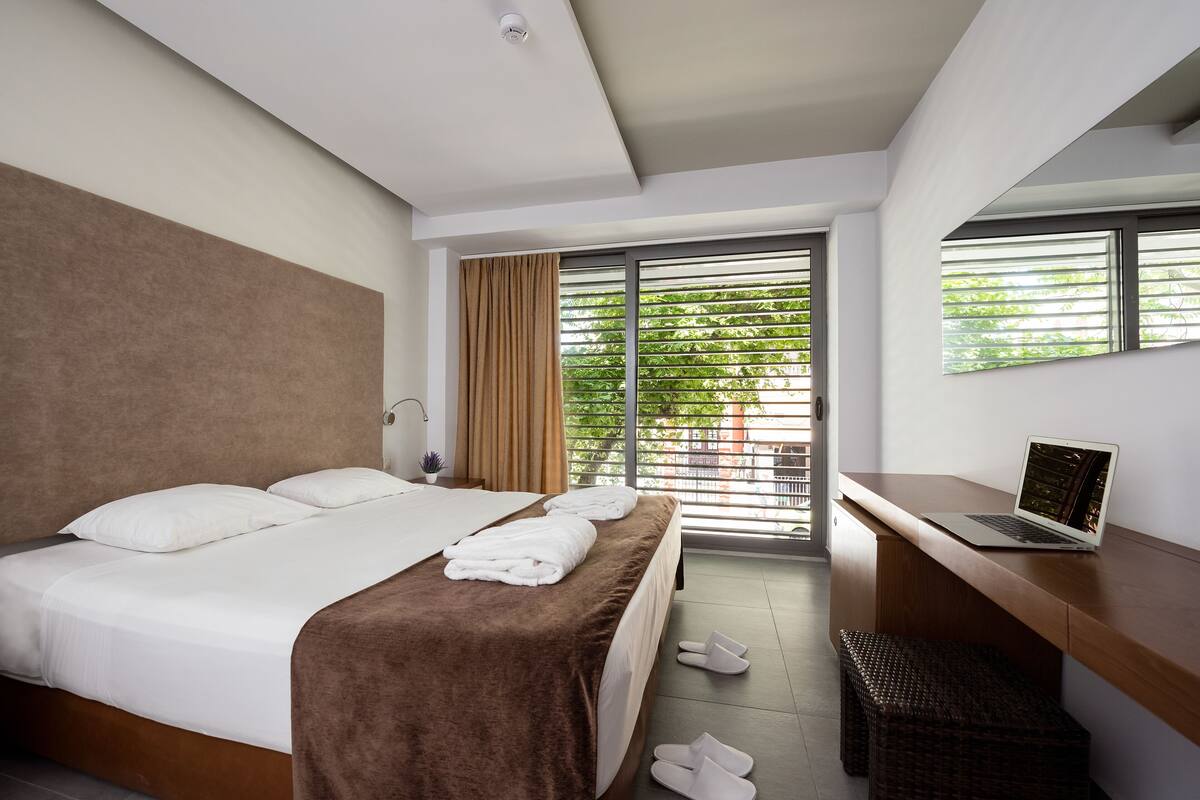 A modern hotel room with a large bed, a desk with a laptop, a window with blinds, and a view of greenery outside.