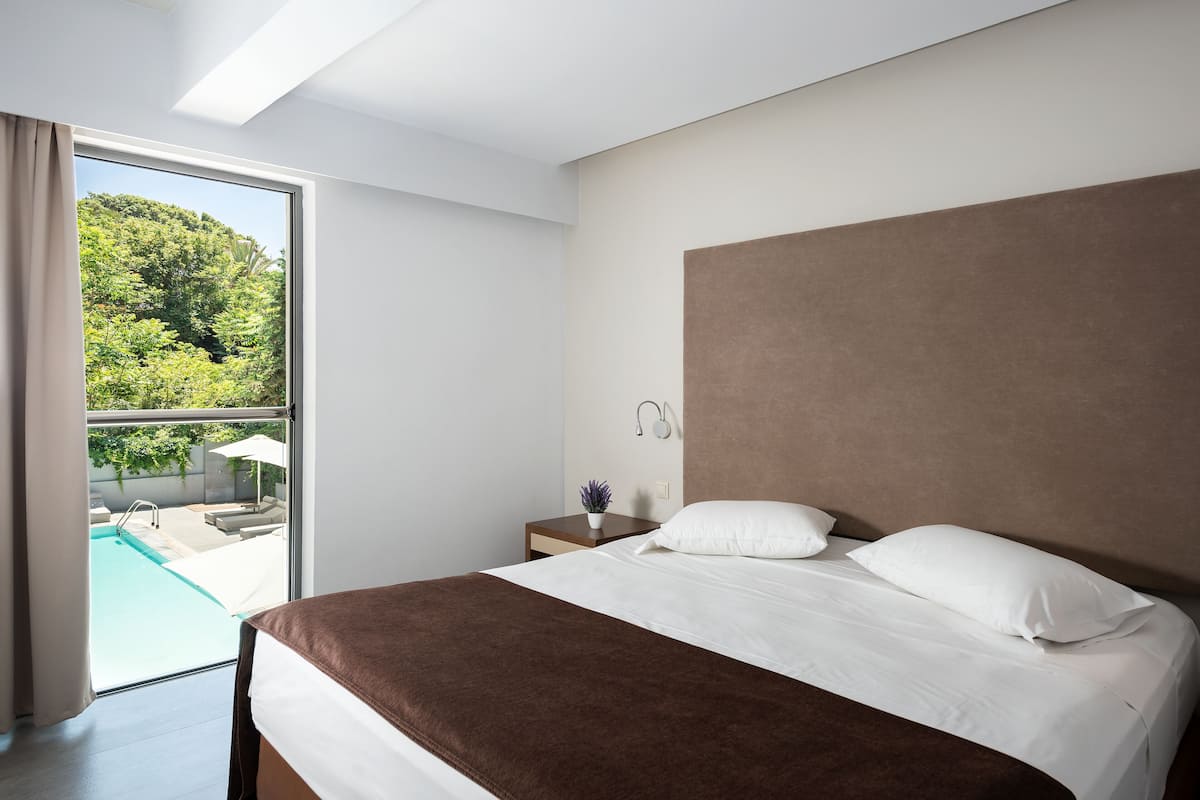 A modern hotel room with a large bed, a nightstand with a lamp, and a view of a swimming pool and greenery outside.