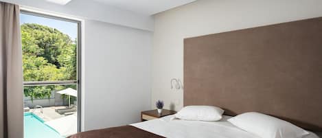 Habitación de hotel moderna con una cama grande, una mesita de noche con lámpara y vistas a una piscina y vegetación.