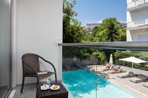 A balcony with a wicker chair, a small table with a plate and a cup, and a view of a swimming pool with lounge chairs and umbrellas. - Oktober Downtown Boutique Hotel (Rhodes)