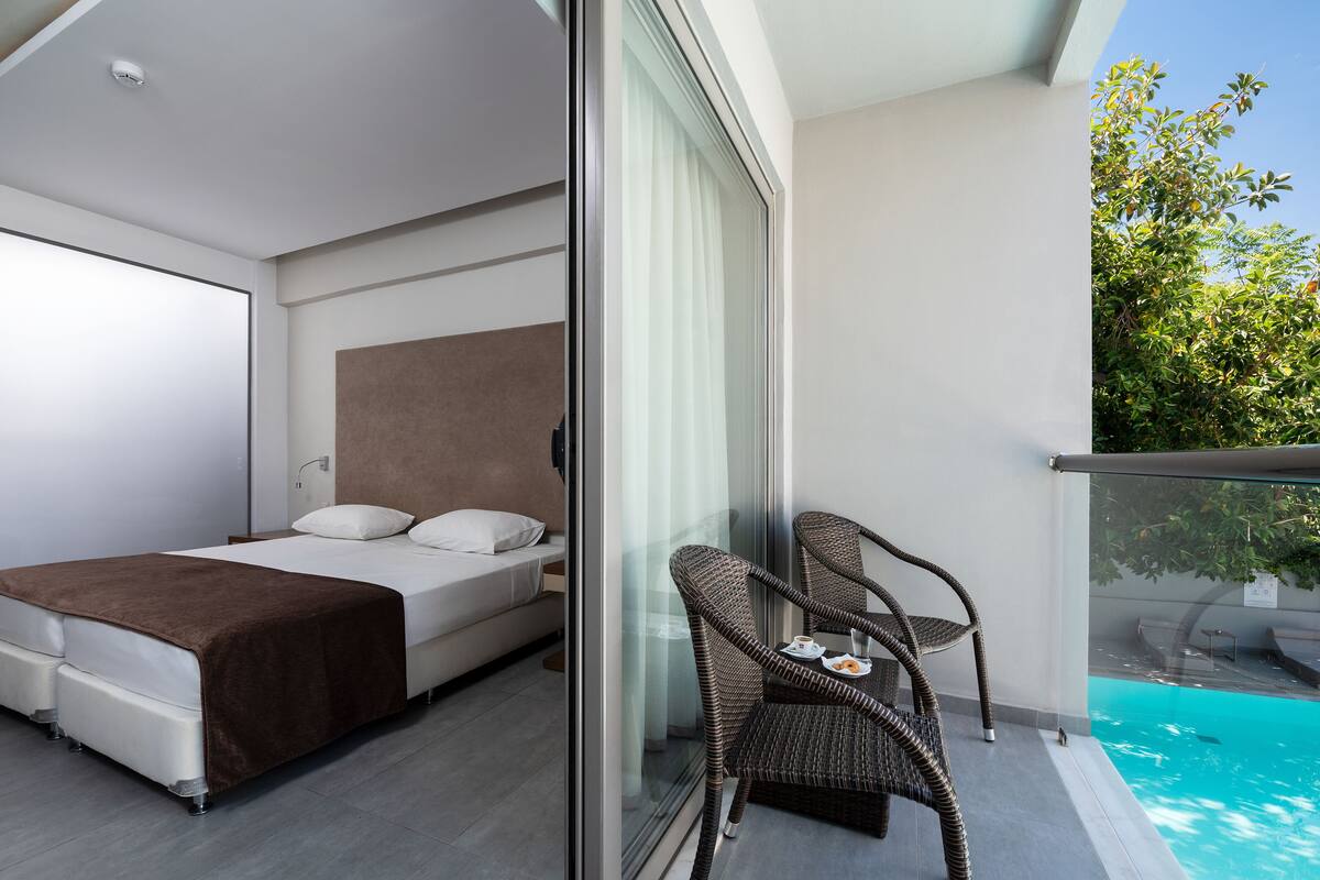 A modern hotel room with a bed, a glass partition, a balcony with wicker chairs, and a view of a swimming pool.