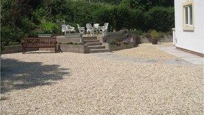 Een tuin met een grindpad, een houten bankje en witte stoelen.