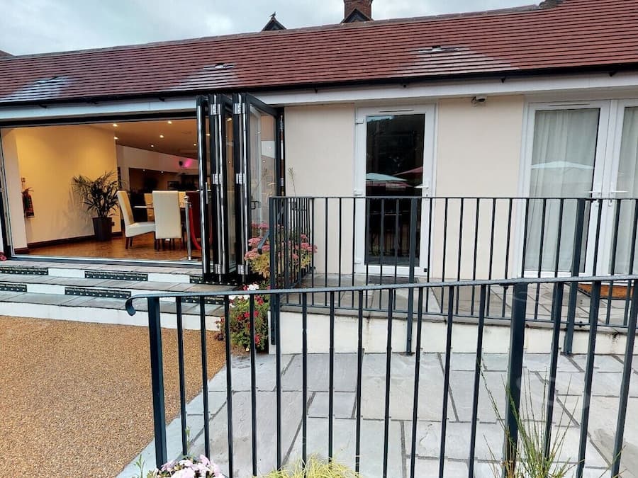 Entrada de un hotel con mostrador de recepción, una escalera que lleva a la entrada y un área exterior cercada con plantas en macetas.