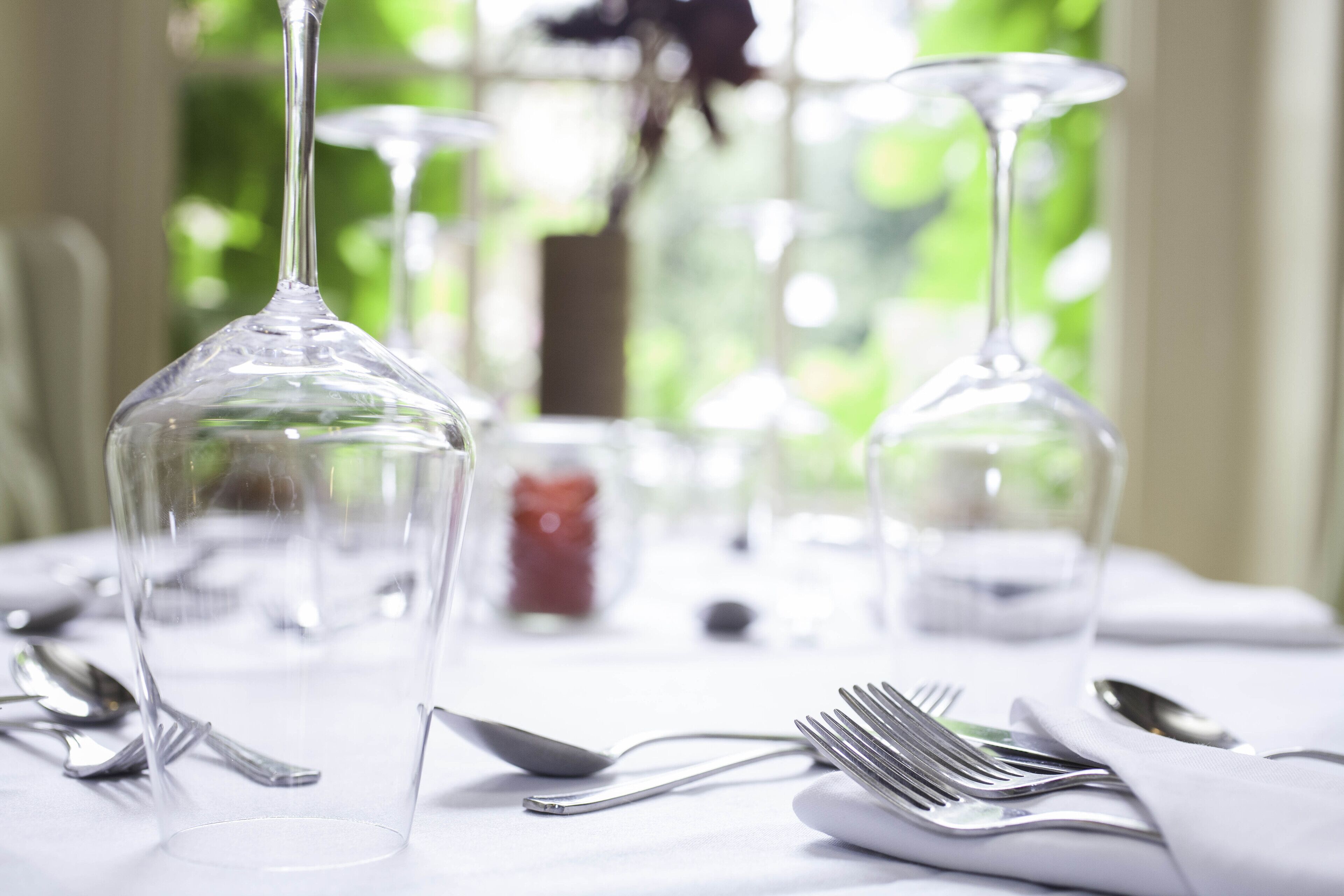 A table set with clear wine glasses, forks, and spoons, with a blurred green background.