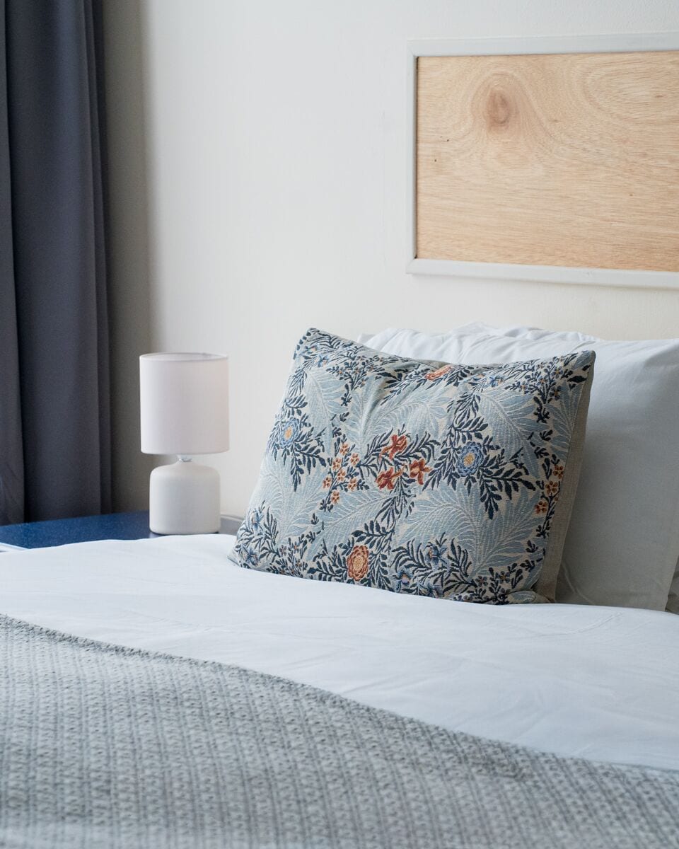 A neatly made bed with a patterned pillow, a white bedside lamp, and a framed picture on the wall.