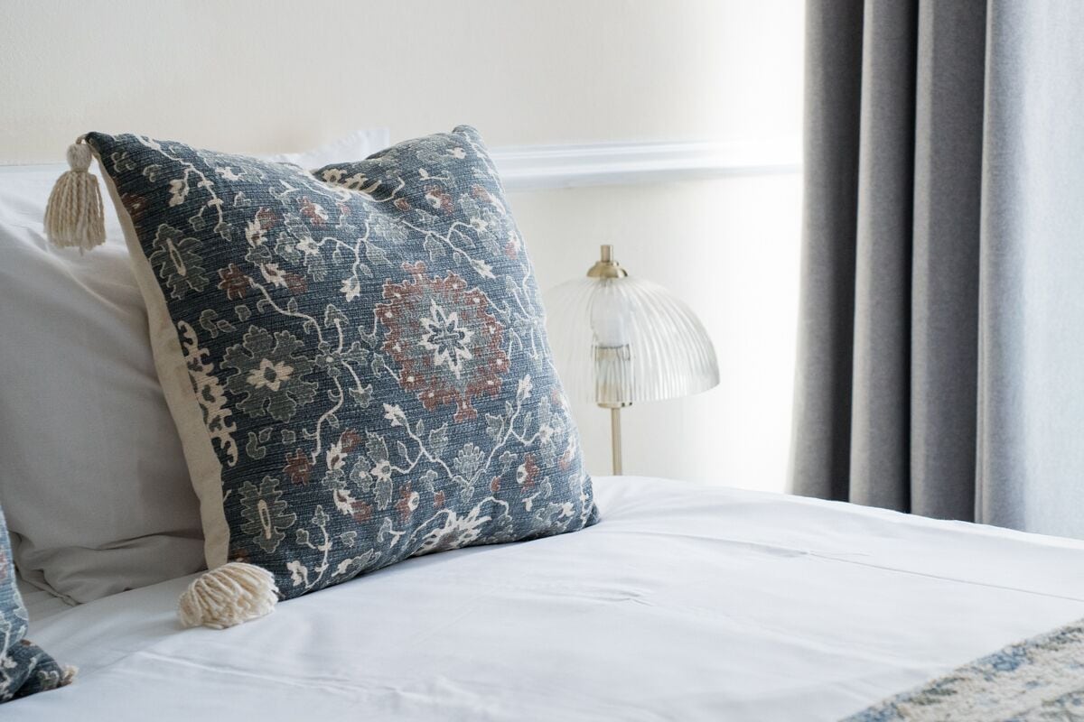 Un lit bien fait, avec un oreiller à motifs, une lampe et des rideaux.