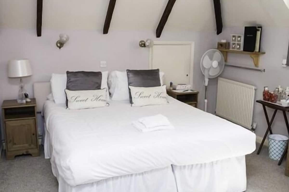 A bedroom with a large bed, two bedside tables, a nightstand, a bedside lamp, a radiator, a small table with a fan, and a shelf with books and decorative items.
