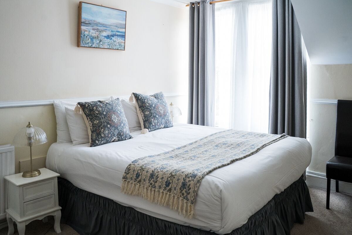 A neatly made bed with a white duvet and dark skirted legs, a bedside lamp, and a framed picture on the wall.