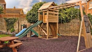 Estructura de madera para juegos con un tobogán y zona de escalada, rodeada por un muro de ladrillo y vegetación.