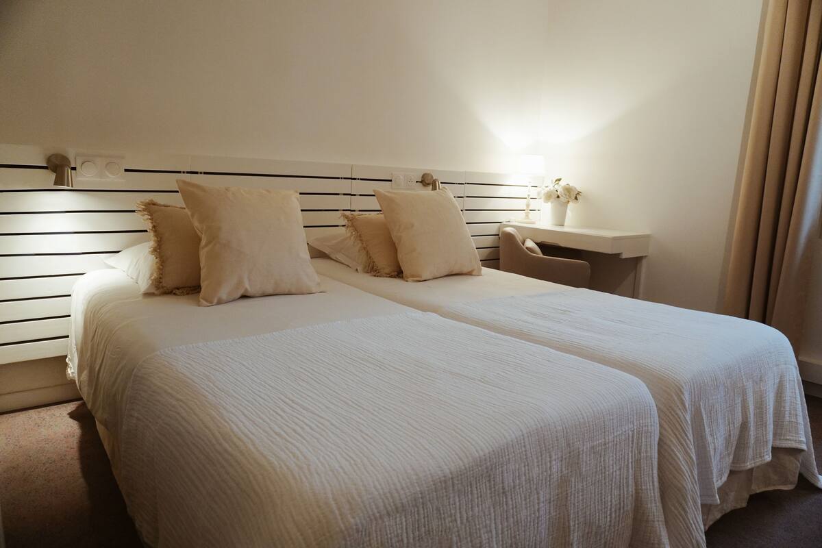 Uma cama de casal com duas almofadas, uma mesa de cabeceira com abajur e um criado-mudo.