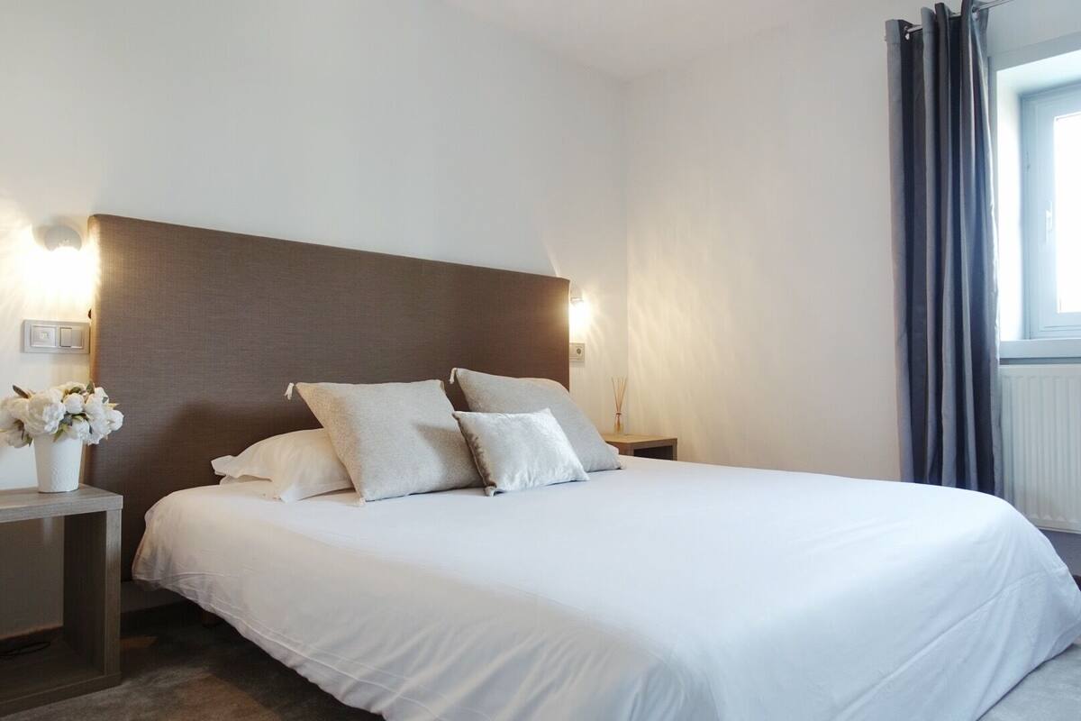 Uma cama bem arrumada com lençóis brancos e duas almofadas, uma mesa de cabeceira com um vaso de flores e um aplique de parede.