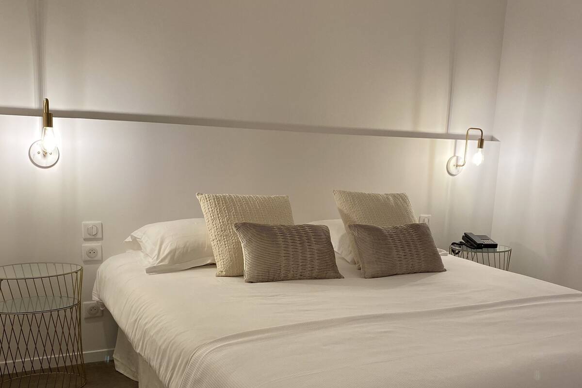 Uma cama bem arrumada com roupa de cama branca e duas almofadas decorativas, uma mesa de cabeceira com abajur, e uma pequena cesta de arame no chão.