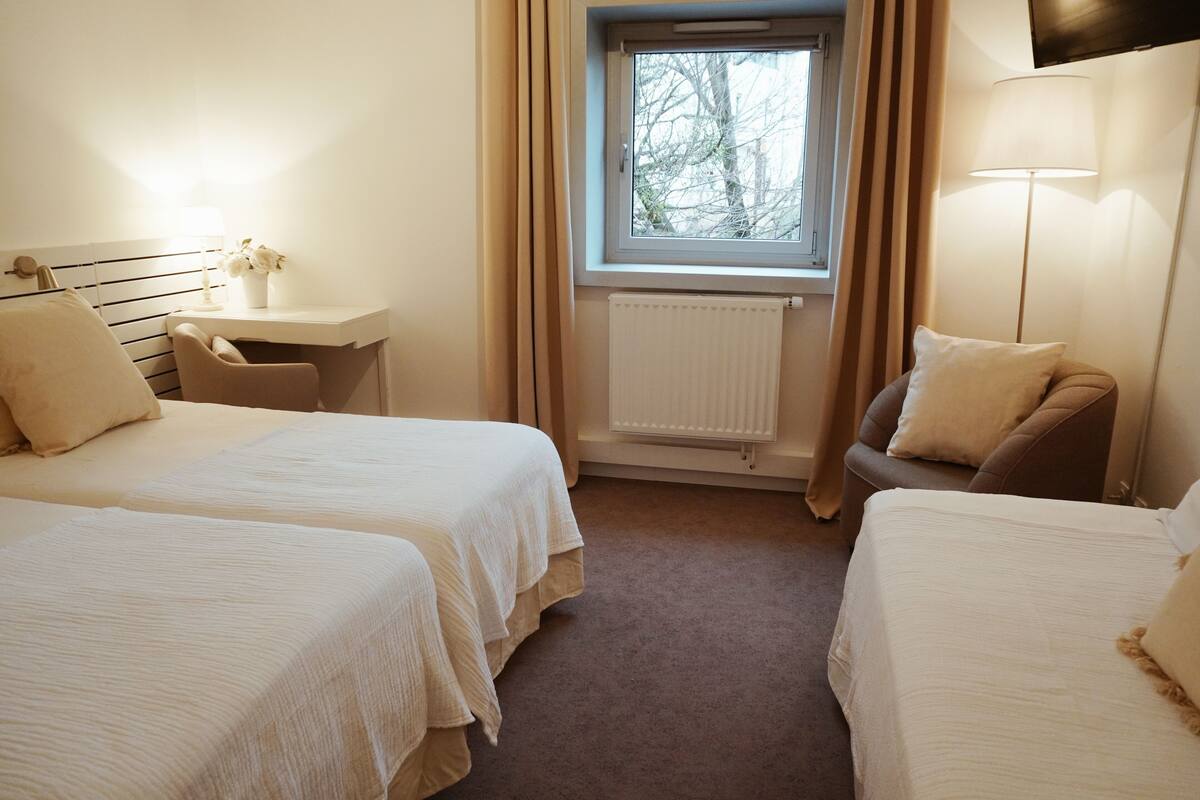Quarto de hotel com duas camas, uma escrivaninha, uma cadeira, janela com cortinas e radiador.