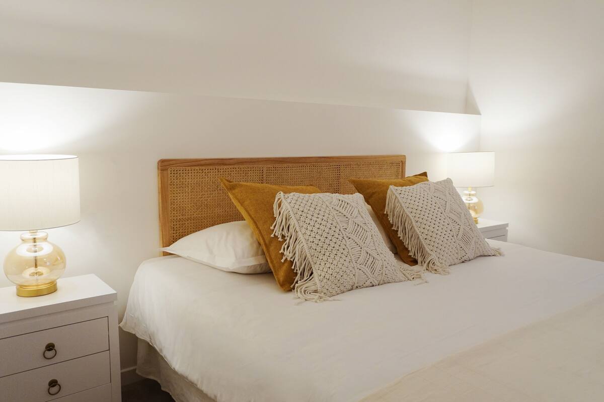 Cama bem arrumada com cabeceira trançada, duas luminárias laterais e mesa de cabeceira branca com gavetas.