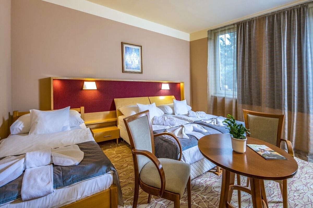 Ein Hotelzimmer mit zwei Betten, einem hölzernen Kopfteil, einem Esstisch mit Stühlen, einer Topfpflanze und einem Fenster mit Vorhängen.