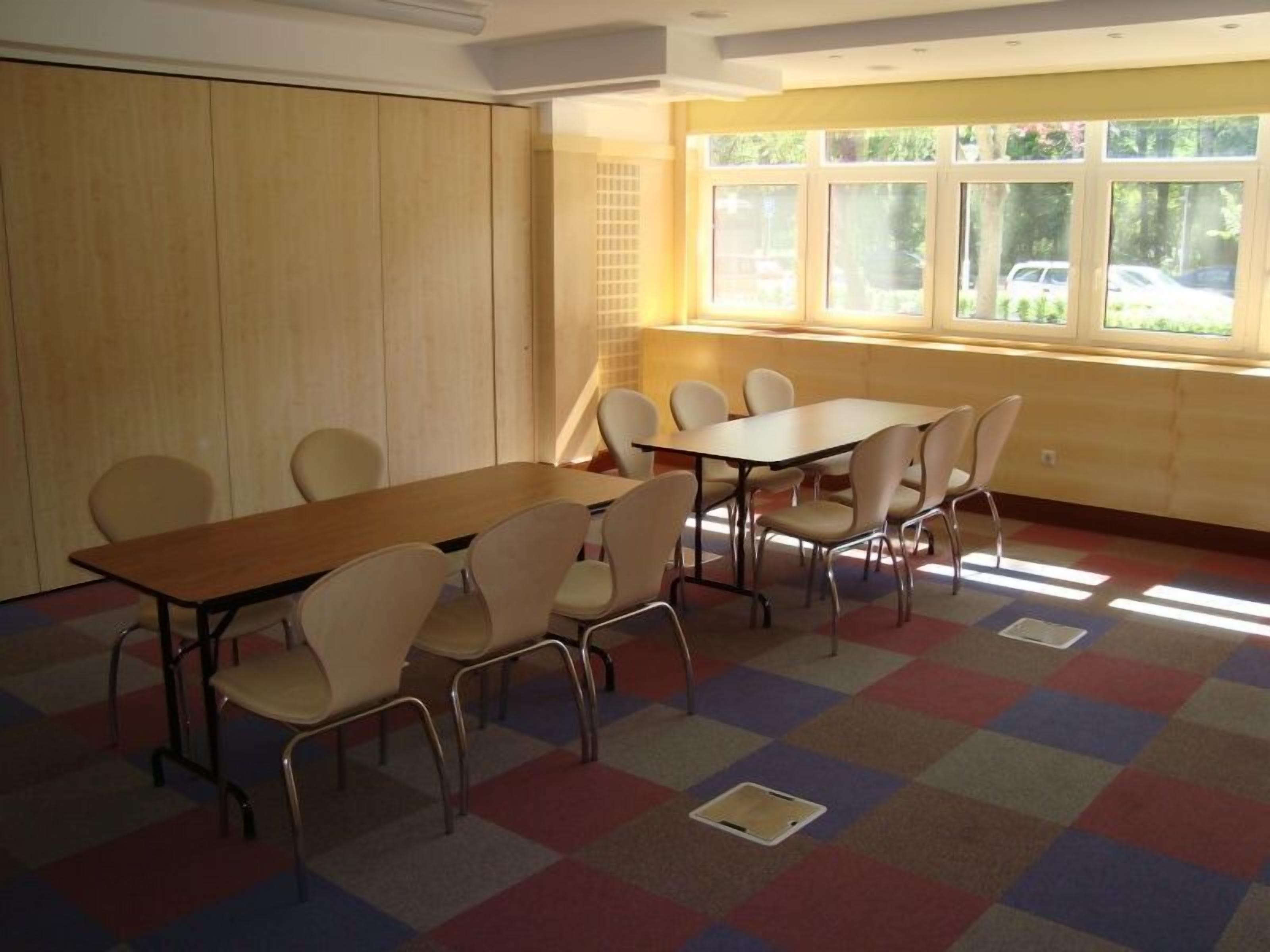 Un salón de conferencias con una larga mesa y sillas, amplias ventanas con vista a coches estacionados y un suelo de baldosas de colores formando un diseño de cuadros.