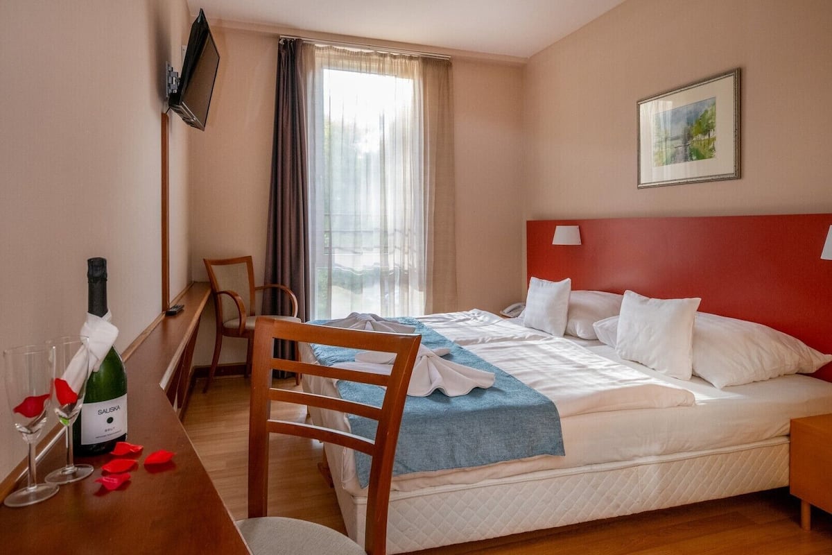 Ein Hotelzimmer mit Bett, Schreibtisch, einer Weinflasche, einem Sessel, einem an der Wand befestigten Fernseher und einem Fenster mit Vorhängen.