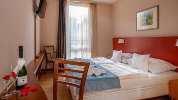 Een hotelkamer met een bed, een bureau met een fles wijn, een stoel, een aan de muur bevestigde televisie en een raam met gordijnen.