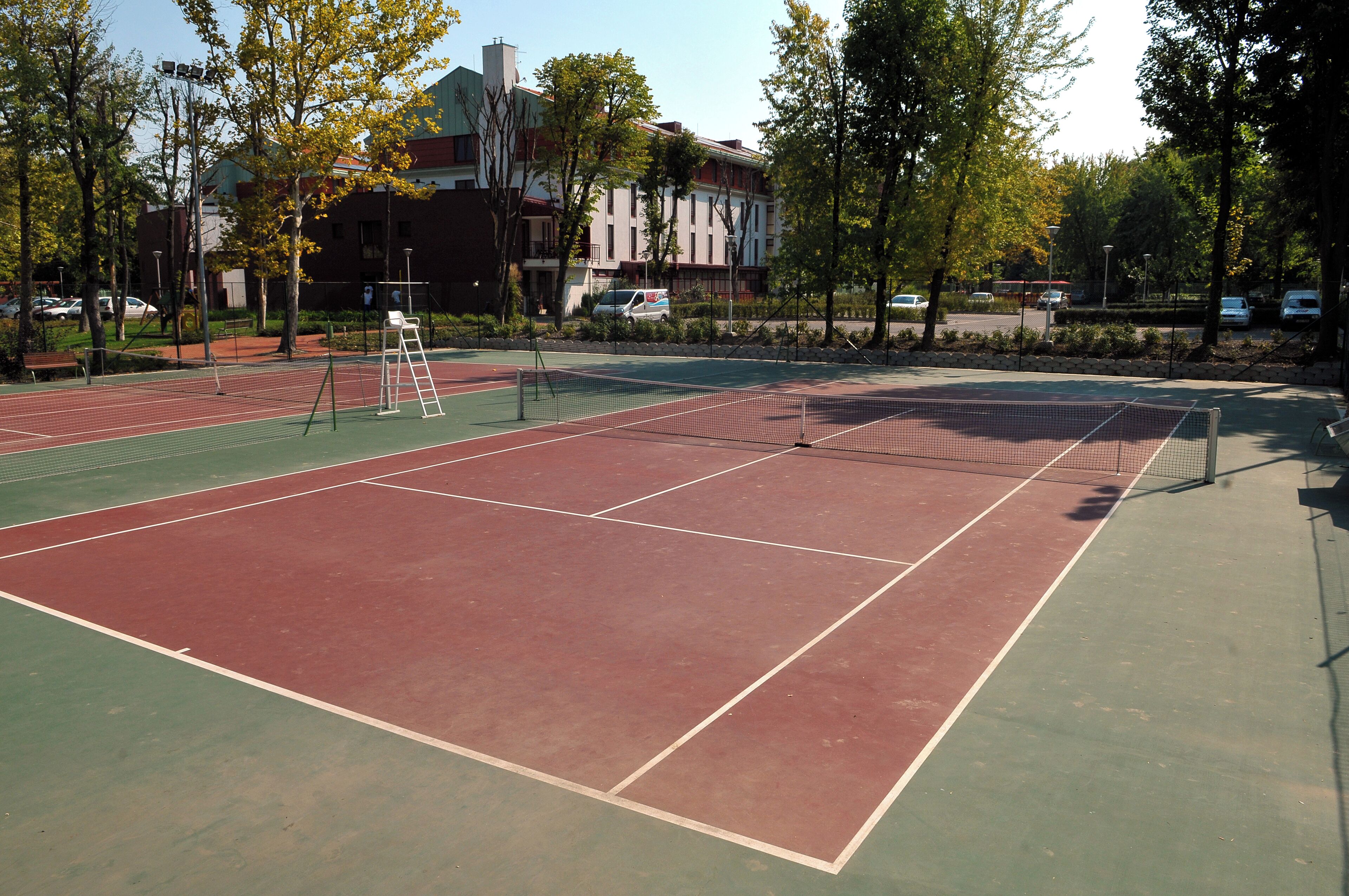 Una cancha de tenis al aire libre con superficie verde y roja, rodeada de árboles y edificios.