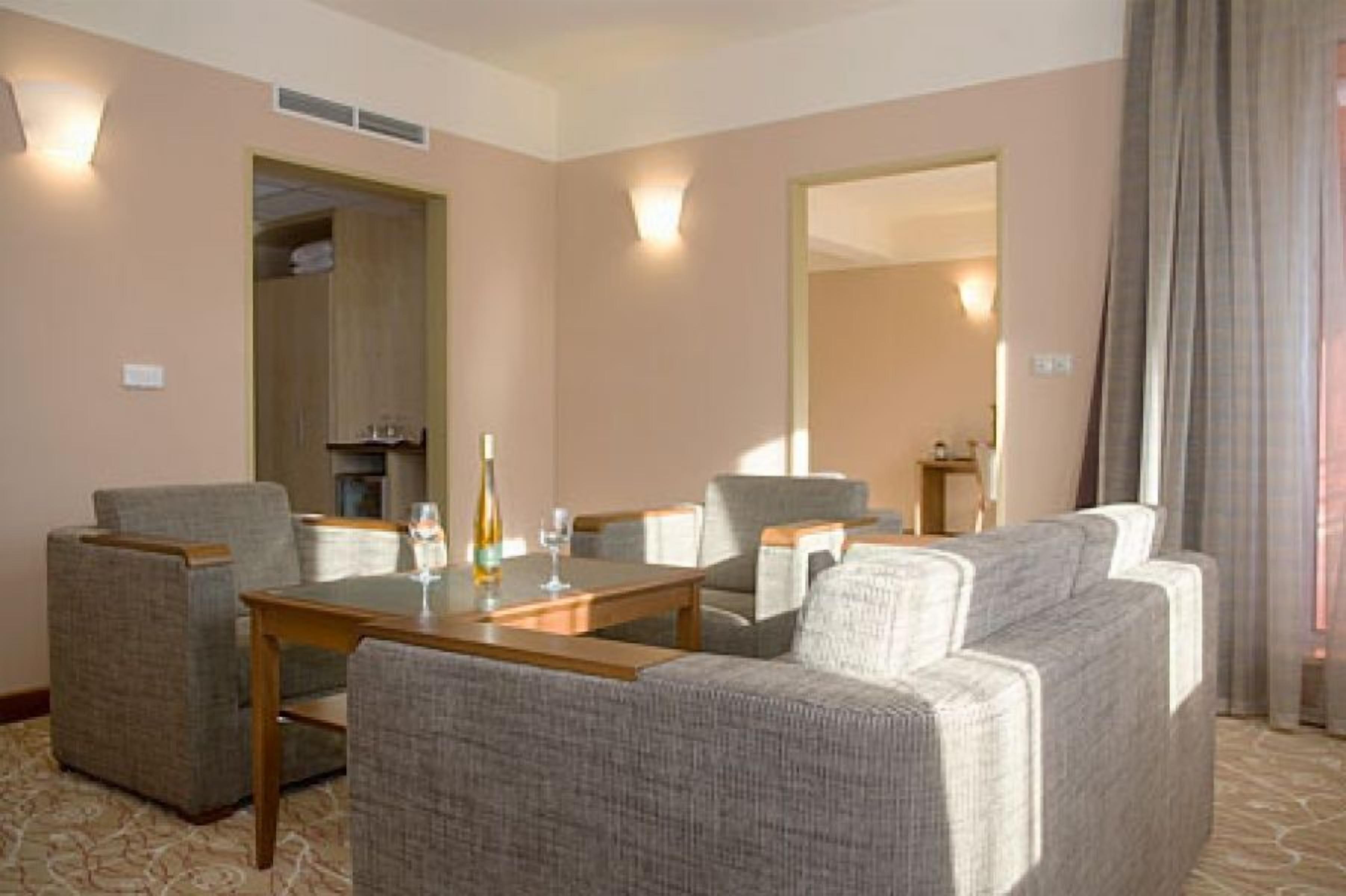 Una habitación de hotel con una mesa de comedor, cuatro sillones, un sofá, un minibar y un espejo grande.