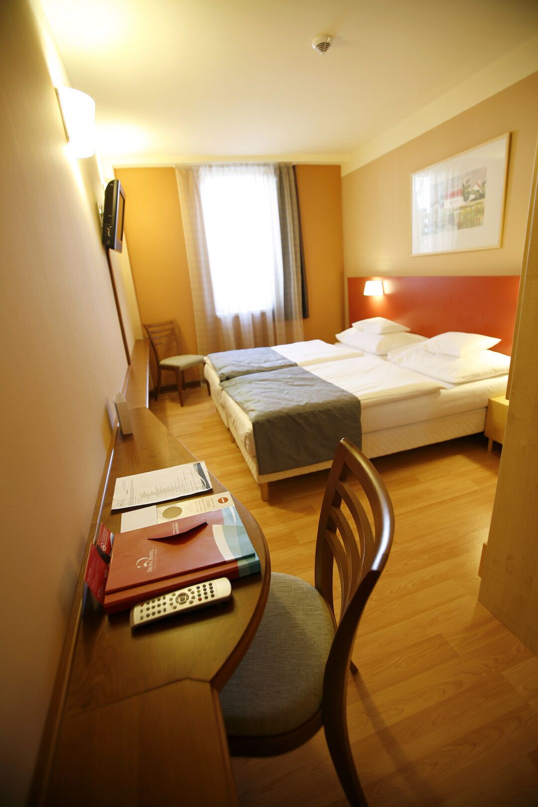 Quarto de hotel com duas camas, uma escrivaninha com cadeira, uma janela com cortinas e uma luminária fixada na parede.