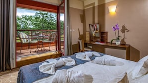 A hotel room with a bed, a desk, a chair, a balcony with chairs, and a view of trees.